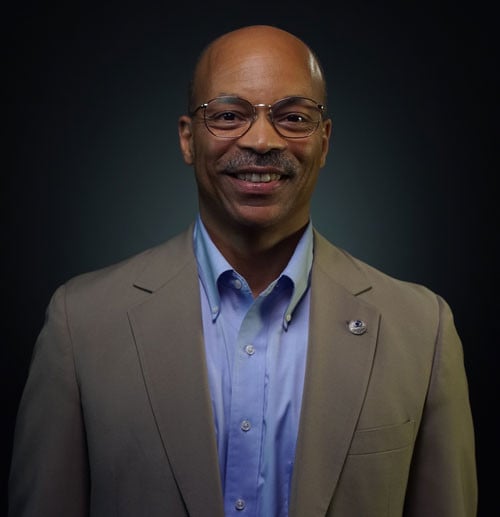 Headshot of Dr. Tom Hogan wearing round glasses a tan suit coat and a light blue dress shirt.