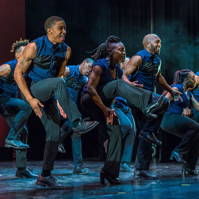 A group of Black dancers wearing blue vest and black pants look joyous while performing a step routine. Most of the group has their right knee raised and are about to clap their hands under their raised legs.